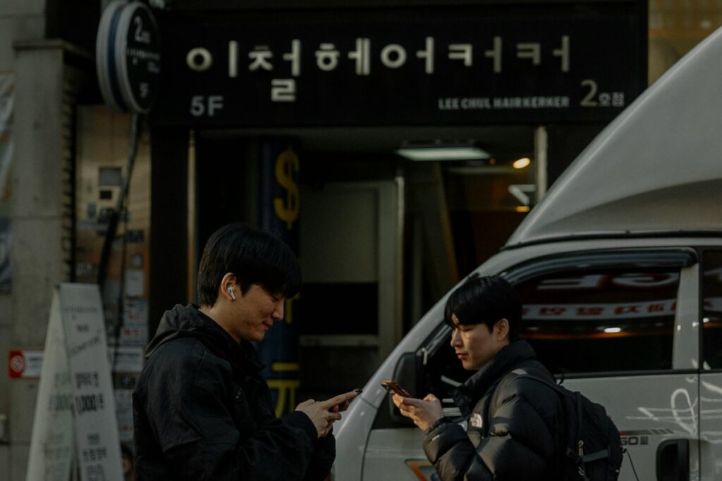 2025 AfreecaTV Marketing Guide for Korea: In an urban corner of Korea, two young men stand engrossed in their phones in front of a building with Korean - language signage. The setting reflects the digital - centric daily life in Korea. For marketers, understanding such real - life scenarios can inspire effective strategies on AfreecaTV, a platform where locals engage with digital content, to reach and resonate with the target audience.