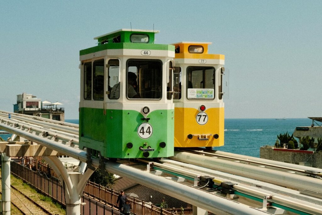 2025 Band Marketing Guide for Korea: Two colorful tram cars, one green numbered 44 and the other yellow numbered 77, run on an elevated track by the sea. The bright hues of the trams contrast with the deep blue ocean in the background. This scenic and unique transportation view can inspire creative marketing approaches on Band in Korea, highlighting the country's blend of modernity and natural beauty to engage local users.