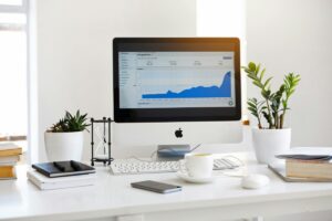 A neatly - arranged workspace features an Apple computer displaying a graph on its screen. Beside it are potted plants, an hourglass, a cup of coffee, and some books. This organized setup might inspire you as you explore the “2025 Douyin Marketing Guide for China”, seeking strategies to thrive on the platform.