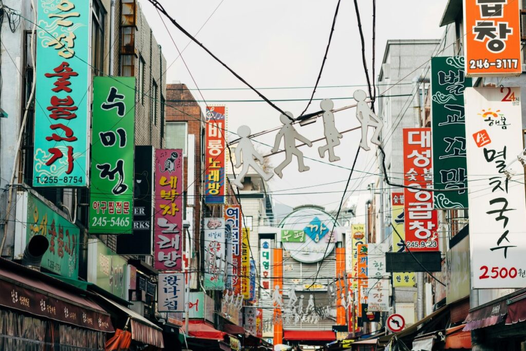 2025 Line Marketing Guide for Korea: A vibrant street scene in Korea, filled with a multitude of colorful signs written in Korean. Hanging above the narrow street are silhouettes of figures, adding a whimsical touch. This bustling urban view showcases the lively commercial atmosphere in Korea, presenting opportunities for creative marketing on Line to connect with local audiences.