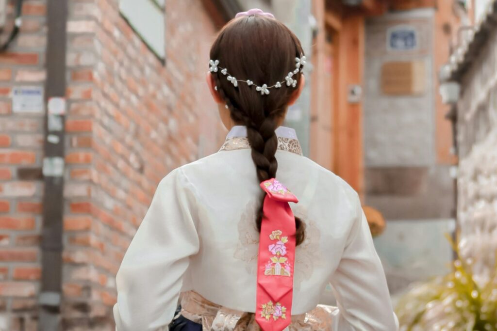 In 2025 Naver Marketing Guide for Korea, a girl in a traditional hanbok with an intricately braided hair adorned by a delicate pearl headpiece walks through a quaint alley. The vibrant pink ribbon with floral patterns on her braid stands out against the muted tones of the brick - lined path. This image captures the essence of Korean traditional beauty, a cultural aspect marketers can leverage on Naver to connect with local audiences.