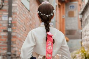 In 2025 Naver Marketing Guide for Korea, a girl in a traditional hanbok with an intricately braided hair adorned by a delicate pearl headpiece walks through a quaint alley. The vibrant pink ribbon with floral patterns on her braid stands out against the muted tones of the brick - lined path. This image captures the essence of Korean traditional beauty, a cultural aspect marketers can leverage on Naver to connect with local audiences.