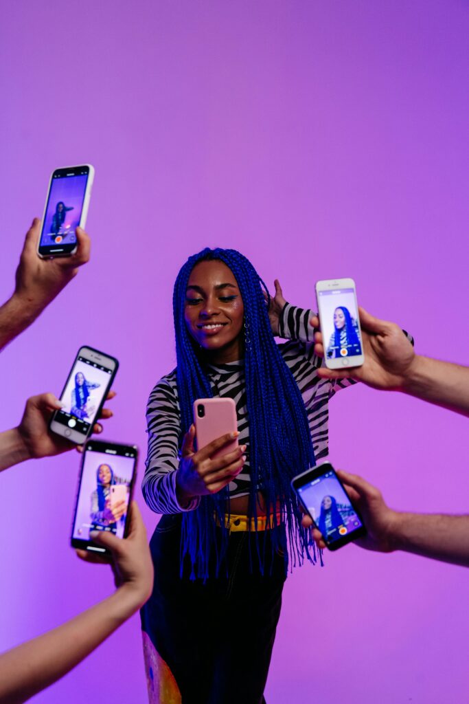 Seorang influencer dengan rambut berwarna biru sedang bersenang - senang sambil diaturi beberapa orang yang memegang ponsel, masing - masing ponsel menampilkan foto atau video influencer itu. Ini menggambarkan dinamika 2025 Tren Pemasaran Influencer Global, di mana kehadiran digital dan interaksi dengan audiens melalui ponsel menjadi penting.