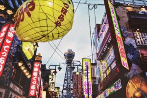 The image captures a vibrant street scene in Japan, with large, colorful lanterns and numerous signs featuring Japanese characters. In the background, a tall tower stands out. This lively urban setting is related to the "2025 Twitter (X) Marketing Guide for Japan", suggesting the dynamic marketing opportunities that exist within Japan's unique cultural landscape on the X platform.