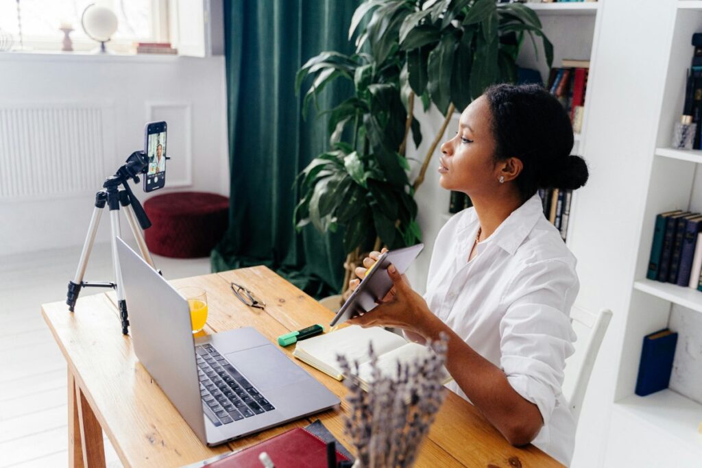 A woman in a white shirt sits at a wooden desk, looking up as she holds a tablet. In front of her, a smartphone on a tripod is recording, and a laptop is open. Around are plants, bookshelves filled with books, and items like a glass of orange juice. This setting could be inspiring as one delves into the "2025 WeChat Marketing Guide for China", exploring ways to succeed in WeChat - based marketing in the Chinese market.