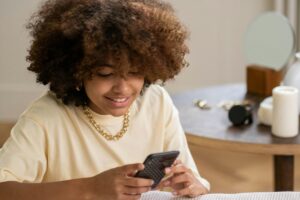 A person with curly hair, wearing a beige top and a gold chain necklace, is smiling while intently looking at a smartphone. In the background, there's a table with a mirror, some accessories, and a candle. This cozy scene might inspire you as you explore the "2025 Xiaohongshu Marketing Guide for China", learning how to make your brand shine on the platform.