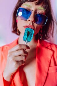鮮やかなオレンジ色のジャケットを着用し、大きなサングラスをかけた人物が、中にカニが封入されたブルーのアイスキャンディを持っています。この画像は、2025年、グローバルインフルエンサーマーケティングのトレンドの中で、個性的で目を引くアイデアや商品が注目を集めることを反映しています。