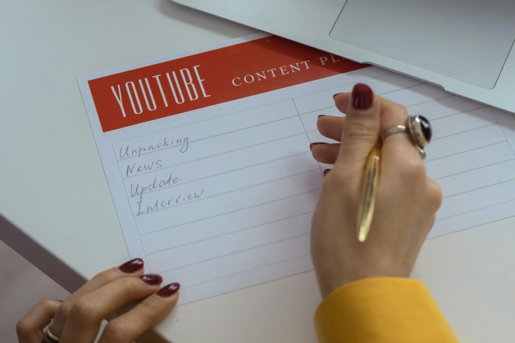 一只涂着深红色指甲油的手拿着金色的笔，正在一张标有“YOUTUBE CONTENT PLAN”的纸上书写，纸上已经写下了“Unpacking”“News”“Update”“Interview”等内容，旁边还放着一台笔记本电脑。这张图片与YouTube内容规划相关，展现了创作者为YouTube平台制定内容计划的场景 。