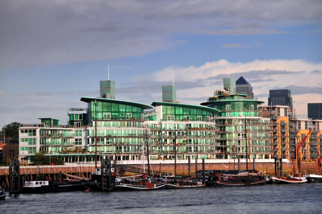 2025年英国Twitter营销百科：河畔，现代感十足的绿玻璃建筑林立，独特的弧形外观与高耸塔楼引人注目。下方停泊着古朴船只，新旧景致交织，展现英国城市独特韵味 。