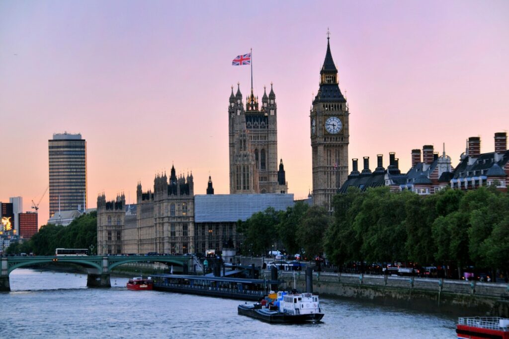 2025年英国YouTube营销秘籍：黄昏时分，泰晤士河畔，标志性的大本钟与威斯敏斯特宫在粉紫色天空下庄严肃穆，飘扬的英国国旗增添一抹亮色，河上船只往来，尽显伦敦独特风情 。
