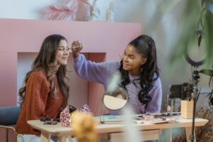 Two women are engaged in a makeup session at a table filled with beauty products. One is applying makeup to the other. In the backdrop, there's a setup suggesting a filming environment. Amidst such creative content creation, an important topic is the Analysis of Market Share in India's Social Media Short Video Platforms.