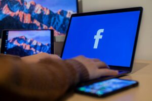 A person's hands are actively typing on a laptop with the Facebook logo displayed on its blue screen. Beside the laptop, there's a smartphone, and in the background, another device shows a scenic mountain image. This setup relates to the context of "Best Platforms for Facebook Influencer Cross - Border Deals", suggesting the exploration or management of cross - border influencer collaborations on Facebook.