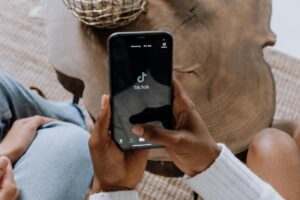 Hands hold a smartphone with the TikTok app open, showing the "Following" and "For You" tabs at the top. The device is set against a backdrop featuring a wooden surface and part of a person's body. This scene relates to "Best TikTok Influencer Cross-Border Collaboration Platforms", hinting at the importance of TikTok in cross - border influencer partnerships.