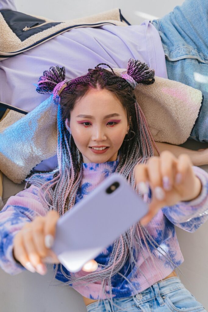 Una joven con trenzas multicolores y una camisa de tie - dye se toma una foto con su teléfono mientras está en un ambiente relajado. Además de esta forma de expresarse personalmente, un tema importante para las empresas es: Cómo aprovechar Xiaohongshu para marcas en China.
