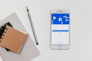 Un teléfono inteligente en una superficie blanca muestra la interfaz de Facebook, con opciones de inicio de sesión y la posibilidad de crear una nueva cuenta. Junto al teléfono hay un bolígrafo y dos carpetas de apuntes. Esta imagen ilustra la interacción cotidiana con la plataforma. Además, si estás interesado en cómo aumentar tus ingresos, descubre "Cómo ganar dinero con FACEBOOK", y aprende a aprovechar las oportunidades que ofrece esta red social.