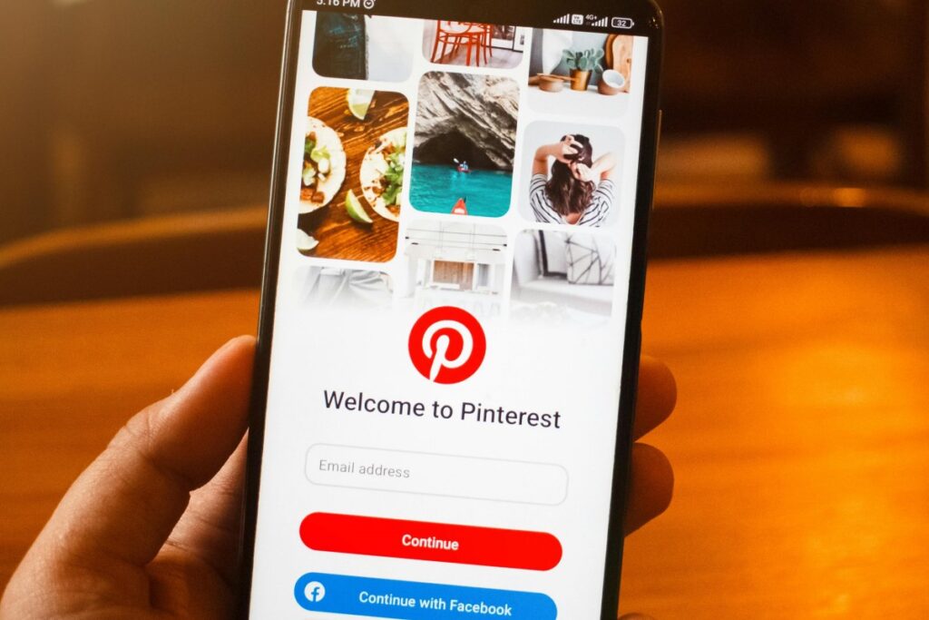 Una mano sostiene un teléfono móvil donde la pantalla muestra la interfaz de bienvenida de Pinterest, con la opción de ingresar una dirección de correo electrónico y botones para continuar o hacerlo con Facebook. Esta imagen se asocia con la guía "Cómo ganar dinero con Pinterest en 2025", brindando una vista previa de la plataforma clave para generar ingresos.