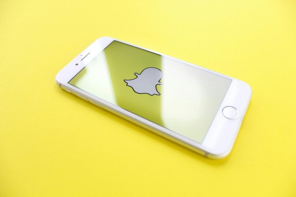 Un teléfono móvil blanco sobre un fondo amarillo brillante. En la pantalla del teléfono está el logotipo de Snapchat. Esta imagen está relacionada con la guía "Cómo ganar dinero con Snapchat en 2025", mostrando la plataforma clave para aquellos interesados en generar ingresos a través de ella.