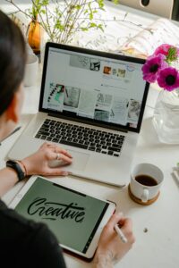 A person is working at a desk, using a laptop with creative - related content on the screen. A tablet in front of the laptop shows the word "Creative". The setting suggests a workspace where strategies for brand promotion might be explored, perhaps related to the topic "Top Bilibili Marketing Strategies to Boost Your Brand in China".