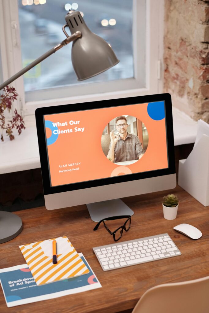 A desktop setup with a computer monitor displaying a "What Our Clients Say" section featuring Alan Mercey, Marketing Head, which is related to global influencer marketing. The desk also has a lamp, a keyboard, a mouse, a pair of glasses, and some stationery items.