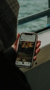Uma pessoa, com uma gorra no cabelo, está segurando um celular com uma capa clara. A tela do celular mostra a interface de uma aplicação de redes sociais, com a seção "Trending" (Em alta) aberta. A página exibe uma publicação de "Lela Abboud", com uma foto de uma paisagem com uma casa e um cão adormecido no chão. No fundo, há uma parede e uma janela com uma vista para o mar.