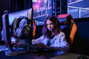Uma mulher de cabelos loiros e ondulados está sentada em uma cadeira gamer nas cores preto e laranja. Ela está concentrada em um computador desktop, com um par de fones de ouvido repousando ao lado do monitor.