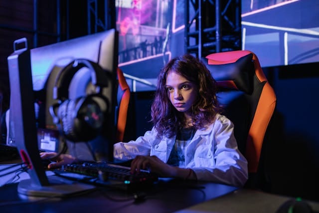 Uma mulher de cabelos loiros e ondulados está sentada em uma cadeira gamer nas cores preto e laranja. Ela está concentrada em um computador desktop, com um par de fones de ouvido repousando ao lado do monitor.