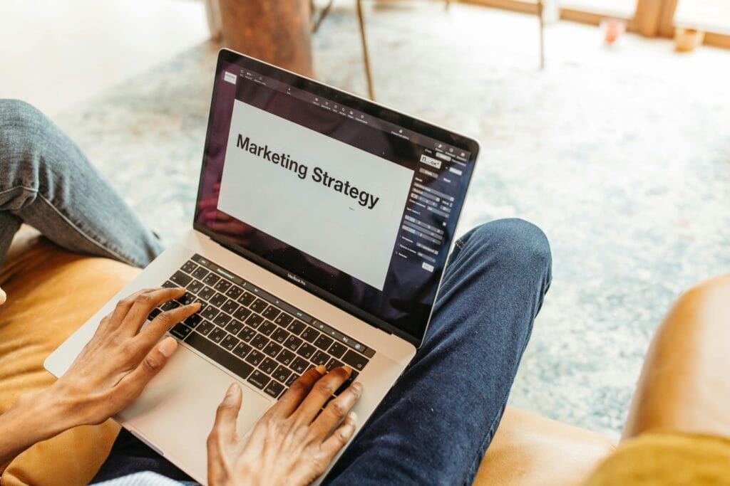 Un individu est assis et a les jambes croisées, tenant un ordinateur portable sur ses genoux. L'écran de l'ordinateur affiche le texte "Marketing Strategy". Les mains de la personne sont posées sur le clavier, comme si elle travaillait sur une stratégie marketing. Le décor du fond semble être un espace de vie lumineux avec des meubles.
