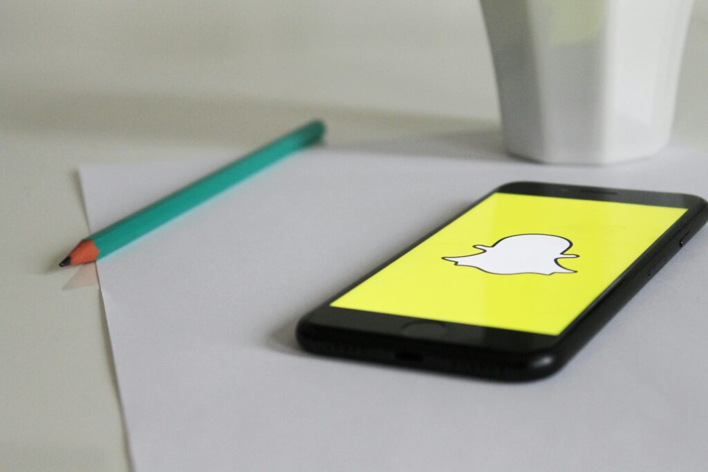 A smartphone on a desk displays the Snapchat logo on its screen. Beside the phone, there is a green pencil and a white cup, suggesting a casual workspace or study area where someone might use Snapchat to communicate and share moments.