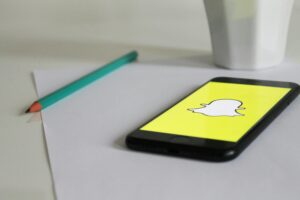 A smartphone on a desk displays the Snapchat logo on its screen. Beside the phone, there is a green pencil and a white cup, suggesting a casual workspace or study area where someone might use Snapchat to communicate and share moments.