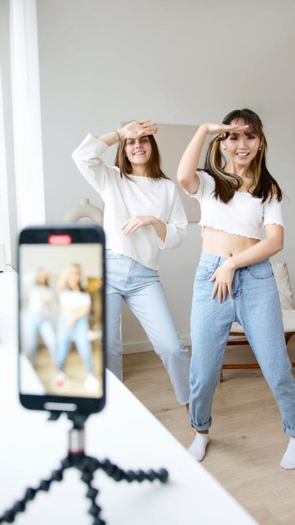 Duas mulheres estão dançando em frente a um celular montado em um tripé, que as filma. Ambas vestem camisetas brancas e calças jeans. Seus rostos irradiam alegria enquanto fazem movimentos com as mãos. O ambiente parece ser um quarto com iluminação natural.