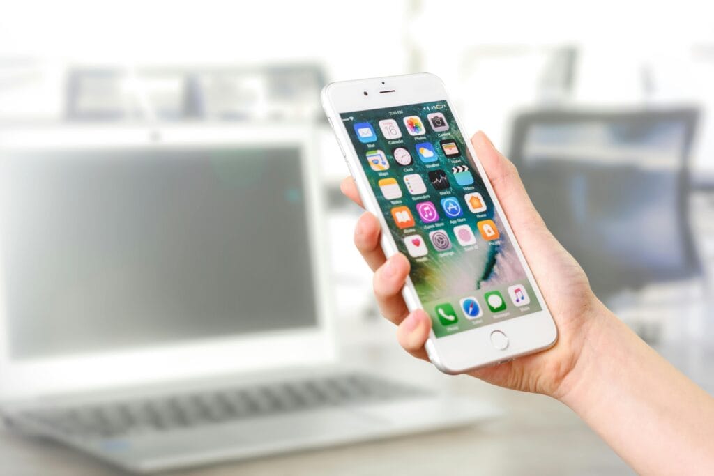 A hand holding a white smartphone with various app icons on the screen, and a laptop in the background.