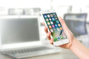 A hand holding a white smartphone with various app icons on the screen, and a laptop in the background.