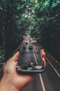 A hand holding a smartphone, with the screen seemingly reflecting a busy road lined with lush green trees.