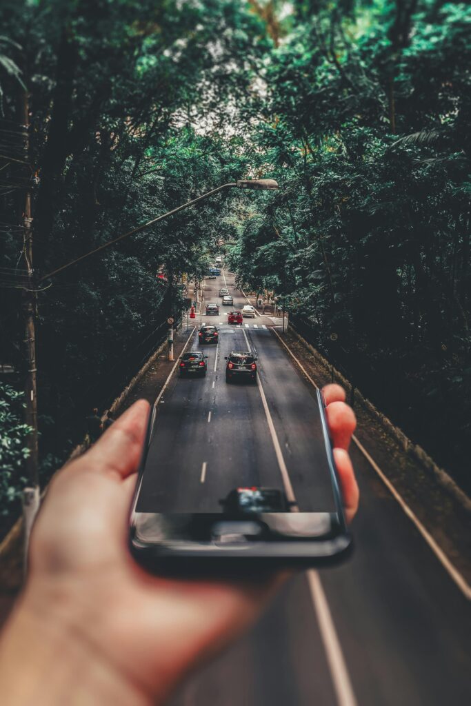 A hand holding a smartphone, with the screen seemingly reflecting a busy road lined with lush green trees.