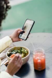 A person holding a smartphone while having a meal that includes a bowl of vegetables and a glass of a red - colored beverage.