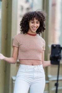 A woman with curly hair, wearing a red - and - white striped cropped top and white jeans, is smiling and gesturing in front of a smartphone on a stand.