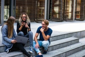 Three people are sitting on steps. One is holding a laptop, another is using a smartphone, and the third is listening attentively.