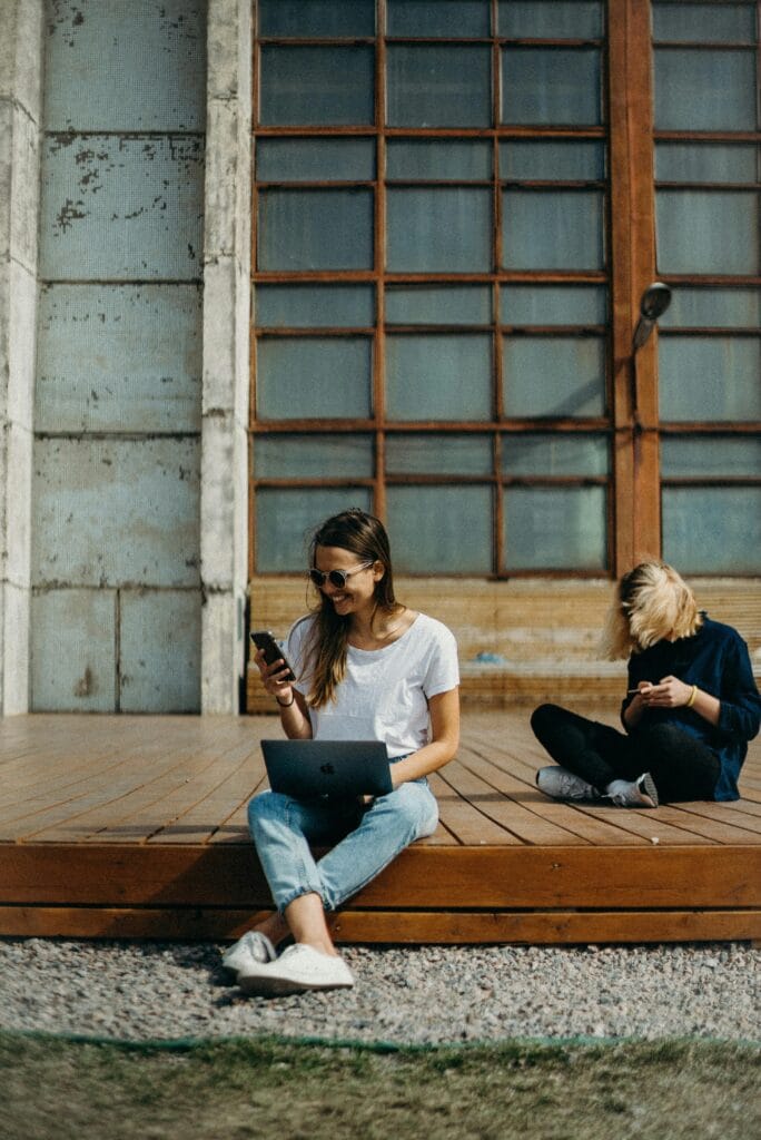 Deux femmes sont assises dehors. L'une est en train d'utiliser un téléphone portable tandis que l'autre utilise un ordinateur portable.