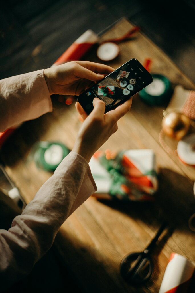 Manos que sostienen un teléfono móvil mientras toman una foto de regalos envueltos en una mesa de madera.