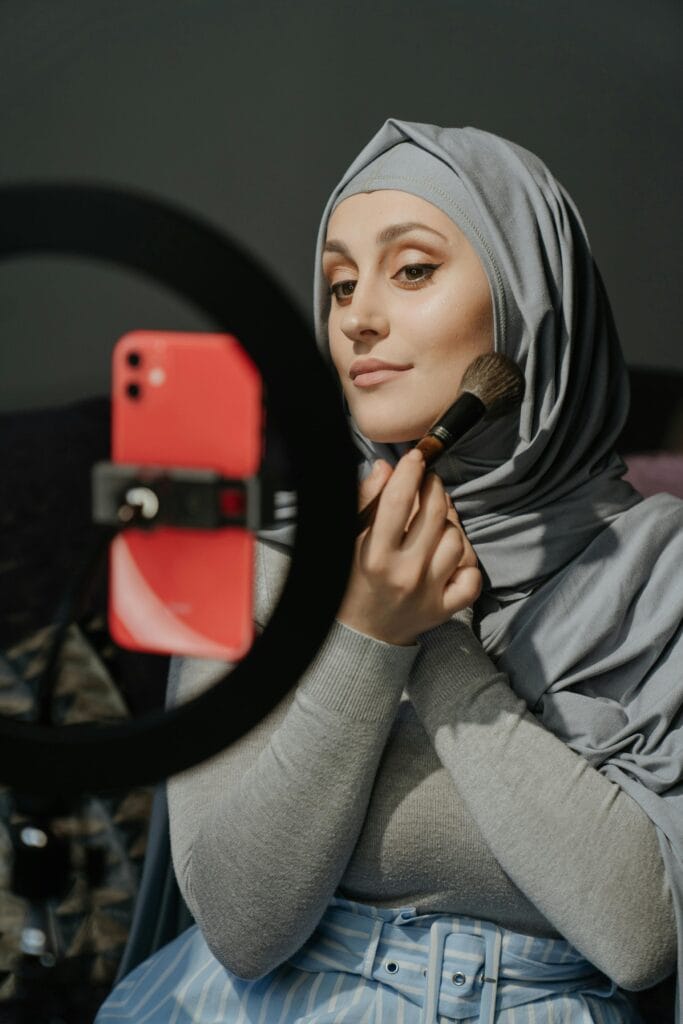 A woman wearing a grey headscarf applies makeup with a brush while a red smartphone is mounted on a ring - light setup.