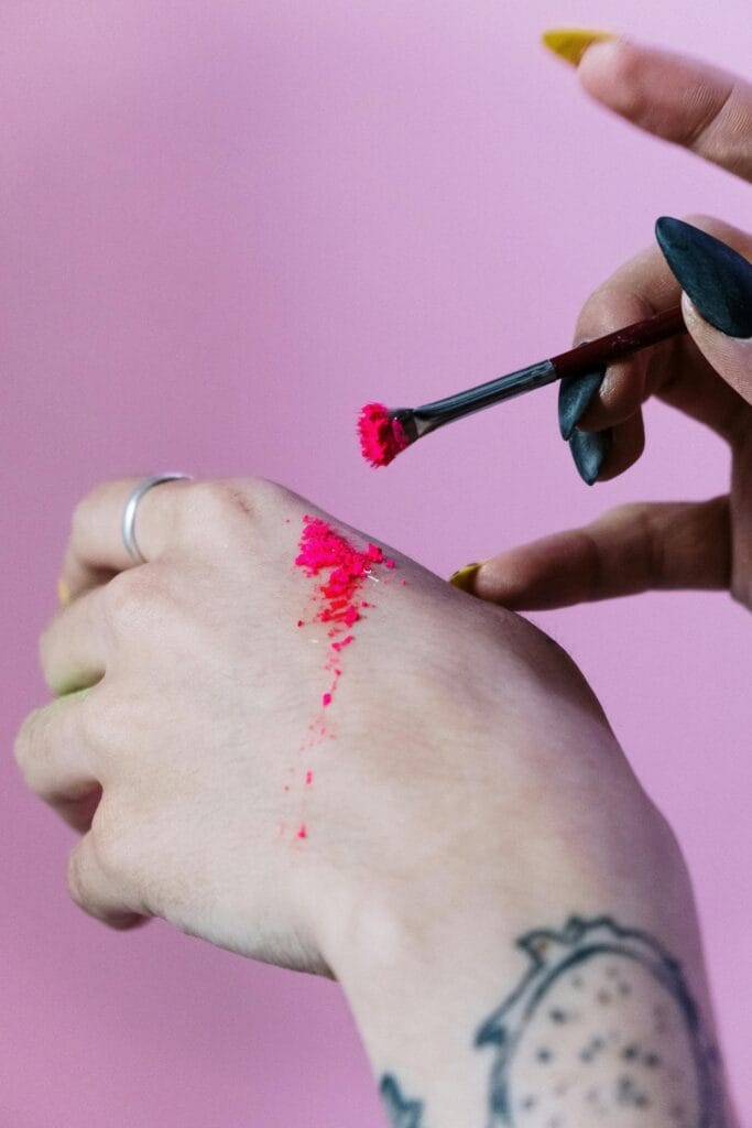 Una mano con un anillo en el dedo, mostrando una tinta de color fucsia aplicada con un pincel, mientras otra mano con uñas pintadas sostiene el pincel. En la mano, también hay una tatuaje. El fondo es de color rosa.