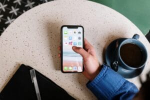 Una mano sostiene un teléfono móvil mostrando la pantalla principal con varios iconos de aplicaciones. Junto al teléfono, hay una taza de café sobre una plato, un bolígrafo y un bloc de notas sobre una mesa con un patrón de terrazzo.