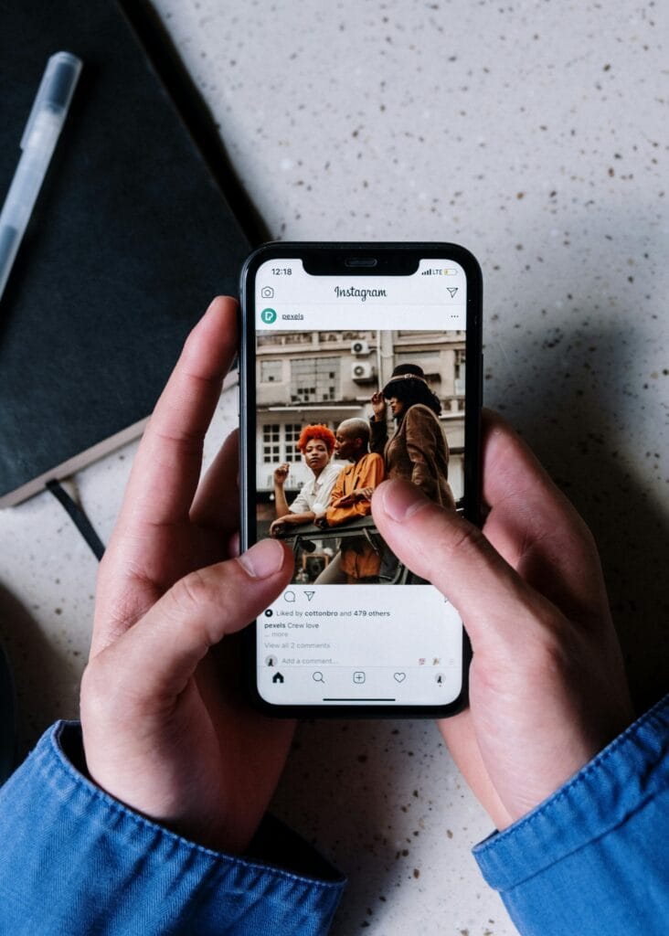 يدان تحمل هاتفًا ذكيًا، تعرض شاشته تطبيق Instagram حيث تظهر صورة لمجموعة من الرجال والنساء مع بعض التفاصيل والتعليقات التفاعلية. في الخلفية، يوجد دفتر مفتوح عليه ملاحظات مكتوبة، مما يضفي لمسة من الإبداع والتنظيم على المشهد.