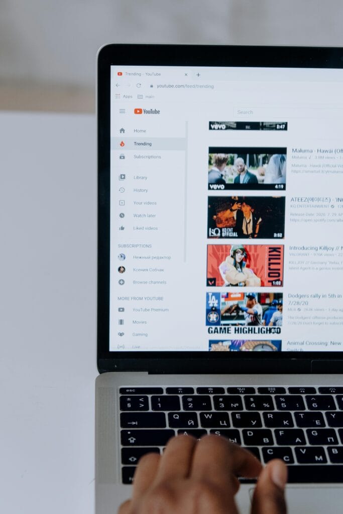 Pantalla de un portátil mostrando la página de "Trending" (Tendencias) en YouTube. En la barra lateral izquierda, se encuentran opciones como "Home" (Inicio), "Trending", "Subscriptions" (Suscripciones), entre otras. A la derecha, se presentan varios thumbnails de vídeos populares, incluyendo canciones de artistas como Maluma y ATEEZ, así como un video relacionado con "Killyjoy" y un resumen de un juego de béisbol de los Dodgers. Una mano está escrita en la teclado del portátil.