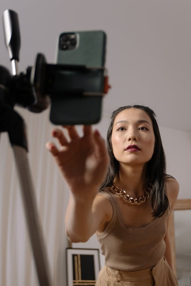 一位女士正伸手调整架在三脚架上的手机，她穿着无袖上衣，戴着一条金色项链，背景中有装饰画等物品。