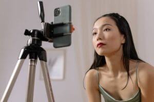 Девушка с длинными, густыми кудрями в стильной белой куртке и синих брюках стоит у окна, задумчиво глядя на экран своего телефона. На дисплее отражается лента социальной сети, а за окном виднеется городской пейзаж с силуэтами зданий.