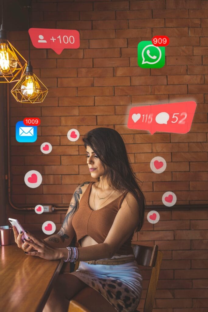 A woman with tattoos on her arm is seated at a wooden table in a room with a brick - wall background and hanging light fixtures. She is focused on her smartphone. Floating around her are digital icons representing social media notifications such as new followers, messages, and likes. xiaohongshu, a popular lifestyle and social media platform, often features similar scenes of users engaging with their content and receiving interactions from the community.