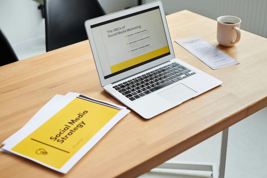 A wooden desk holds an open laptop displaying a presentation titled "The ABCs of Social Media Marketing." Beside the laptop are printed materials with the title "Social Media Strategy" and a beige coffee cup. Weixin, a powerful social media and communication platform, is an important aspect of social media marketing strategies like the ones being explored in this setup.