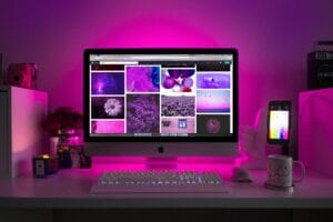 An Apple iMac on a desk is illuminated by vibrant pink ambient lighting. The screen displays a grid of colorful images. Surrounding the computer are various items including a smartphone, a coffee - mug with a pattern, a small plant, and some decorative objects. Kuaishou, a popular short - video platform, could be a great source for finding content similar to the visually appealing images on the screen, whether it's for inspiration in photography or design.