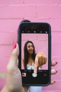 A hand with bright red - painted nails is holding a smartphone, with the camera app open. On the screen of the phone, a woman is pointing forward, and the background is a pink brick wall. Douyin, a popular short - video platform, often features such moments of content creation, where users capture engaging and shareable videos like the one being recorded here.
